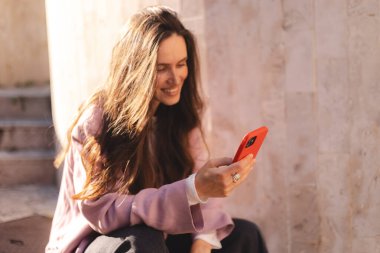 Pembe ceketli gülümseyen genç kadın taş basamaklarda oturuyor, akıllı telefon kullanıyor, mesajlaşıyor ve gülüyor. Rahat duruş, bacak bacak üstüne atma, güneşli günün tadını çıkarma..