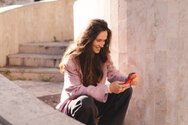 Pembe ceketli gülümseyen genç kadın taş basamaklarda oturuyor, akıllı telefon kullanıyor, mesajlaşıyor ve gülüyor. Rahat duruş, bacak bacak üstüne atma, güneşli günün tadını çıkarma..