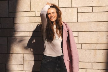 Harika uzun dalgalı esmer, saçları düzelten, saçları düzelten, haftasonunu dışarıda geçiren, omuzlarında pembe ceket olan, beyaz uzun kollu, altın kollu bir kolye takan bir kadının fotoğrafı.. 