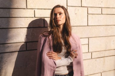 Pembe ceketli, modern taş bir duvara karşı çapraz kollarla poz veren kendinden emin genç bir kadın, sıcak güneş ışığında duruyor. Doğal hafif moda portresi, şık bir duruşu ve rahat duruşu var..