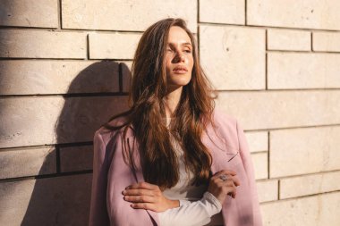 Pembe ceketli, modern taş bir duvara karşı çapraz kollarla poz veren kendinden emin genç bir kadın, sıcak güneş ışığında duruyor. Doğal hafif moda portresi, şık bir duruşu ve rahat duruşu var..
