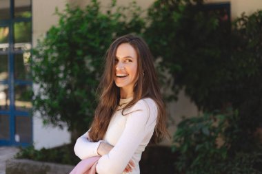 Genç esmer, uzun saçlı iş kadınının kendine güvenen portresi sokak ortasında çapraz ellerle duruyor. Başarılı, gülümseyen bir kadın takım elbisesiyle mutlu ve güler..