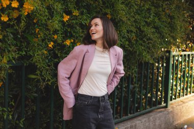 Açık havada güneşli bir günün tadını çıkaran pembe büyük spor ceketli, gülümseyen genç bir kadın. Kendine güvenen, neşeli ve şık, yemyeşil bir çiçeğe karşı duran. Doğal ışık yaşam tarzı anı.