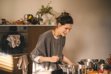 Güzel, mutlu genç bir kadın ev mutfağında yemek pişiriyor ve ocağın tavasındaki çorbayı test ediyor. Mutfakta yemek pişiren kadın tencerede karıştırıyor akşam yemeği hazırlıyor. Genç ev hanımı gülümsüyor.