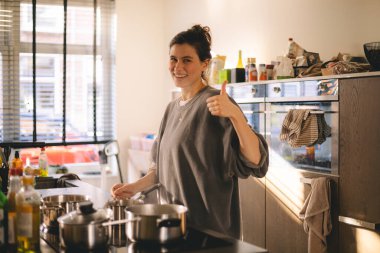 Genç ev hanımı parmaklarını iyi donanımlı, modern bir mutfakta kaldırıyor. Kız tencereli elektrikli sobanın yanında duruyor. Kahvaltı ya da akşam yemeği hazırla..