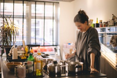 Güzel, mutlu genç bir kadın ev mutfağında yemek pişiriyor ve ocağın tavasındaki çorbayı test ediyor. Mutfakta yemek pişiren kadın tencerede karıştırıyor akşam yemeği hazırlıyor. Genç ev hanımı gülümsüyor.