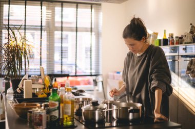 Güzel, mutlu genç bir kadın ev mutfağında yemek pişiriyor ve ocağın tavasındaki çorbayı test ediyor. Mutfakta yemek pişiren kadın tencerede karıştırıyor akşam yemeği hazırlıyor. Genç ev hanımı gülümsüyor.