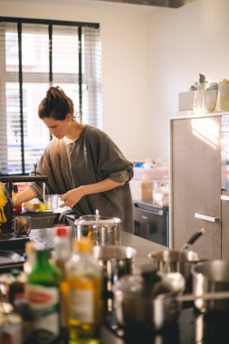 Genç bir kadın, doğal ışık ve günlük malzemelerle dolu rahat bir mutfakta yemek pişiriyor. Gerçek hayata, sadeliğe ve sağlıklı rutine odaklanarak evde yemek hazırlamak için samimi bir yaşam tarzı..
