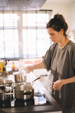 Esmer yüksek çörekli kadın akşam yemeği, öğle yemeği ya da kahvaltı hazırlıyor bardaktan tencereye kadar bulaşıkla su dolduruyor. Genç ev hanımı.