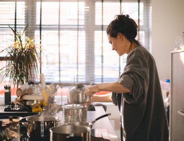 Sıcak bir mutfakta yemek pişiren genç bir kadın sobanın üstünde sıcak bir tencere açarken işine odaklanmış. Doğal ışık, günlük giysiler, sıcak ev atmosferi, günlük hayattan gerçek anlar..