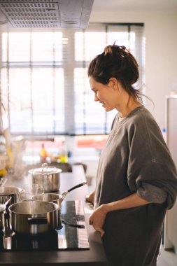 Güzel, mutlu genç bir kadın ev mutfağında yemek pişiriyor ve ocağın tavasındaki çorbayı test ediyor. Mutfakta yemek pişiren kadın tencerede karıştırıyor akşam yemeği hazırlıyor. Genç ev hanımı gülümsüyor.