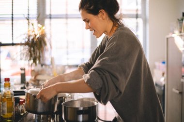 Sıcak bir mutfakta yemek pişiren genç bir kadın sobanın üstünde sıcak bir tencere açarken işine odaklanmış. Doğal ışık, günlük giysiler, sıcak ev atmosferi, günlük hayattan gerçek anlar..