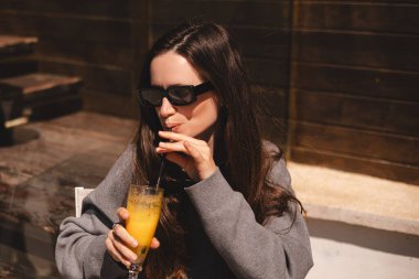 Kafede oturup portakallı limonata, taze meyve suyu ya da kokteyl içen uzun saçlı mutlu esmer. Susuzluğunu giderir. Rahatlama konsepti. Gülen mutlu kadın soğuk kokteyl içiyor..
