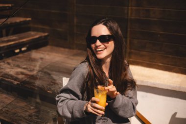 Kafede oturup portakallı limonata, taze meyve suyu ya da kokteyl içen uzun saçlı mutlu esmer. Susuzluğunu giderir. Rahatlama konsepti. Gülen mutlu kadın soğuk kokteyl içiyor..