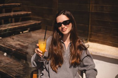 Kafede oturup portakallı limonata, taze meyve suyu ya da kokteyl içen uzun saçlı mutlu esmer. Susuzluğunu giderir. Rahatlama konsepti. Gülen mutlu kadın soğuk kokteyl içiyor..