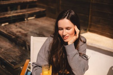 Kahvede oturup portakallı limonata, taze meyve suyu ya da kokteyl içen uzun saçlı esmer kız. Susuzluğunu giderir. Rahatlama konsepti. Gülen mutlu kadın soğuk kokteyl içiyor..