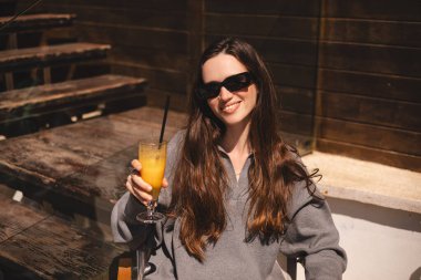 Kafede oturup portakallı limonata, taze meyve suyu ya da kokteyl içen uzun saçlı mutlu esmer. Susuzluğunu giderir. Rahatlama konsepti. Gülen mutlu kadın soğuk kokteyl içiyor..