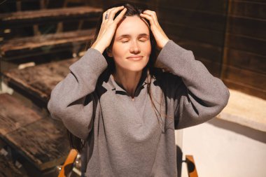 Şirin esmer, uzun saçlı, gülümseyen, saçını düzelten, gözlerini kapatan bir kız. Kadın saçını düzeltiyor. Gri kazak giyiyor, kafede dinleniyor. Mutlu esmer kız flört ediyor ve cilveleşiyor..