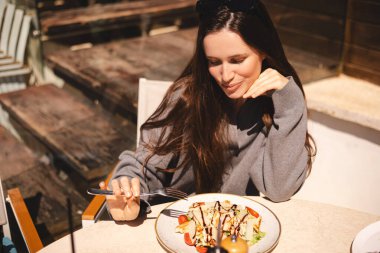 Kafede bir tabak sezar salatasıyla oturan neşeli genç kadın, tavuk ve vişneli domates yemeye hazır. Esmer, uzun saçlı kadın çatallı, tavuklu yemekten hoşlanıyor..