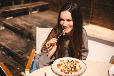 Kafede bir tabak sezar salatasıyla oturan neşeli genç kadın, tavuk ve vişneli domates yemeye hazır. Esmer, uzun saçlı kadın çatallı, tavuklu yemekten hoşlanıyor..