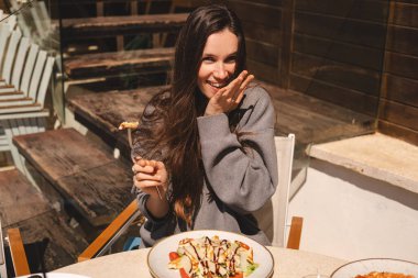 Kafede bir tabak sezar salatasıyla oturan neşeli genç kadın, tavuk ve vişneli domates yemeye hazır. Esmer, uzun saçlı kadın çatallı, tavuklu yemekten hoşlanıyor..