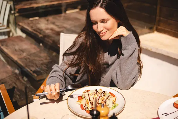 Kafede bir tabak sezar salatasıyla oturan neşeli genç kadın, tavuk ve vişneli domates yemeye hazır. Esmer, uzun saçlı kadın çatallı, tavuklu yemekten hoşlanıyor..