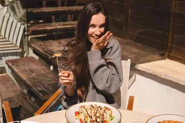 Kafede bir tabak sezar salatasıyla oturan neşeli genç kadın, tavuk ve vişneli domates yemeye hazır. Esmer, uzun saçlı kadın çatallı, tavuklu yemekten hoşlanıyor..