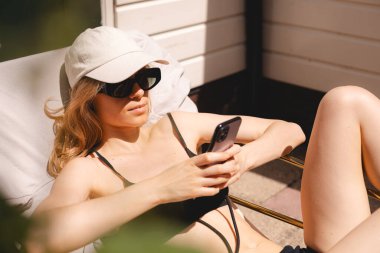 Sakin sarışın kadın arka kapıda güneşleniyor, güneş gözlüğü takıyor, şapka takıyor ve mayo giyiyor. Kadın mesajlaşması, mesaj yazması, haberleri okuması, internet üzerinden cep telefonu kullanması..