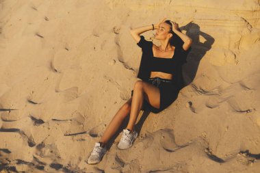 Kısa esmer saçlı, kumların üzerinde oturan ve doğa üzerine profesyonel fotoğraf çekilirken rüya gören çarpıcı bir genç kadın. Dışarıda poz veren siyah gömlekli kadın model yüzünü tut..