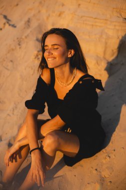 Siyah tişörtlü, kısa saçlı, genç, güzel esmer kadın gün batımında kumların üzerinde oturuyor. Kız rahat ve mutludur. Düşünceli kız dinleniyor, dudaklarını ısırıyor..