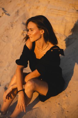 Siyah tişörtlü, kısa saçlı, genç, güzel esmer kadın gün batımında kumların üzerinde oturuyor. Kız rahat ve mutludur. Düşünceli kız dinleniyor, dudaklarını ısırıyor..