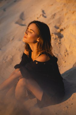 Siyah tişörtlü, kısa saçlı, genç, güzel esmer kadın gün batımında kumların üzerinde oturuyor. Kız rahat ve mutludur. Düşünceli kız dinleniyor..
