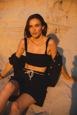 Siyah tişörtlü, kısa saçlı, genç, güzel esmer kadın gün batımında kumların üzerinde oturuyor. Kız rahat ve mutludur. Düşünceli kız dinleniyor..