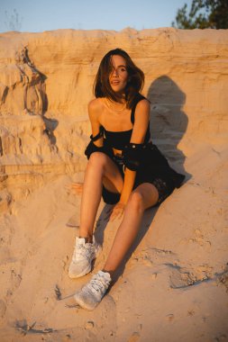 Siyah tişörtlü, kısa saçlı, genç, güzel esmer kadın gün batımında kumların üzerinde oturuyor. Kız rahat ve mutludur. Düşünceli kız dinleniyor..