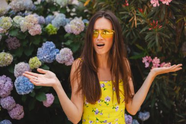 What problem wtf. Pissed-off frustrated and angry young brunette caucasian girl shrugging with confused annoyed expression, cant understand what happened, standing near blossom hydrangea outdoors.