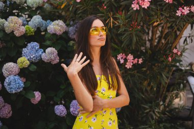 Esmer, uzun saçlı bir kadın sıcak hava, sıcak yaz günü, dudak bükme yüzünden elini sallıyor. Sarı elbiseli ve güneş gözlüklü kız. 