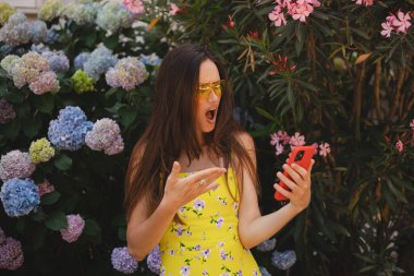 Çiçek fidanlığının yanında cep telefonu kullanan çılgın ve şok edici genç esmer kadın. Mutsuz kadının cep telefonu var. Hayal kırıklığına uğramış üzgün bayan, haberleri dehşete düşürdü. Bu da ne? Olamaz..