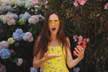 Çiçek fidanlığının yanında cep telefonu kullanan çılgın ve şok edici genç esmer kadın. Mutsuz kadının cep telefonu var. Hayal kırıklığına uğramış üzgün bayan, haberleri dehşete düşürdü. Bu da ne? Olamaz..