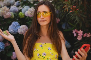 Çiçek fidanlığının yanında cep telefonu kullanan çılgın ve şok edici genç esmer kadın. Mutsuz kadının cep telefonu var. Hayal kırıklığına uğramış üzgün bayan, haberleri dehşete düşürdü. Bu da ne? Olamaz..