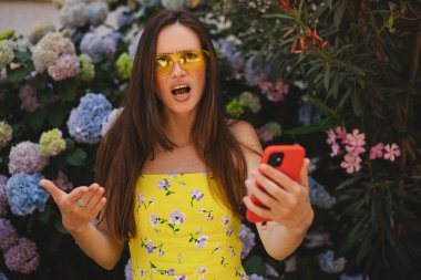 Çiçek fidanlığının yanında cep telefonu kullanan çılgın ve şok edici genç esmer kadın. Mutsuz kadının cep telefonu var. Hayal kırıklığına uğramış üzgün bayan, haberleri dehşete düşürdü. Bu da ne? Olamaz..