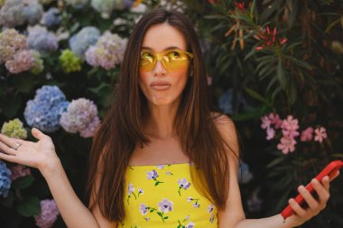 Çiçek fidanlığının yanında cep telefonu kullanan çılgın ve şok edici genç esmer kadın. Mutsuz kadının cep telefonu var. Hayal kırıklığına uğramış üzgün bayan, haberleri dehşete düşürdü. Bu da ne? Olamaz..