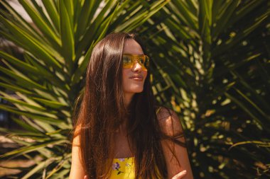 Tropik palmiye ağacının önünde duran gülümseyen genç kadın. Güneşli bir yaz gününde sarı güneş gözlüğü ve çiçekli bir elbise giymek. Neşeli açık hava portresi. Mutlu gülen kız.