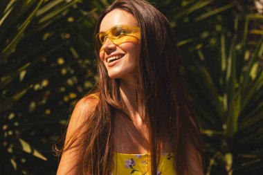 Tropik palmiye ağacının önünde duran gülümseyen genç kadın. Güneşli bir yaz gününde sarı güneş gözlüğü ve çiçekli bir elbise giymek. Neşeli açık hava portresi. Mutlu gülen kız.