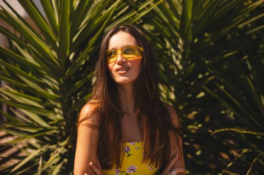 Tropik palmiye ağacının önünde duran gülümseyen genç kadın. Güneşli bir yaz gününde sarı güneş gözlüğü ve çiçekli bir elbise giymek. Neşeli açık hava portresi. Mutlu gülen kız.