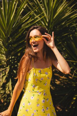 Gülen kadın tropik palmiye ağacının önünde güneş gözlüklerine dokunuyor. Güneşli bir yaz gününde sarı güneş gözlüğü ve çiçekli bir elbise giymek. Neşeli açık hava portresi, vay canına. Duygular yana bakıyor, ağız açık..
