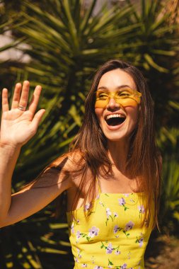 Gülümseyen genç kadın selam veriyor elini tropik palmiye ağacının önünde sallıyor. Güneşli bir yaz gününde sarı güneş gözlüğü ve çiçekli bir elbise giymek. Neşeli açık hava portresi.