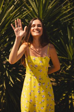 Gülümseyen genç kadın selam veriyor elini tropik palmiye ağacının önünde sallıyor. Güneşli bir yaz gününde sarı çiçekli elbise giymek. Neşeli açık hava portresi.
