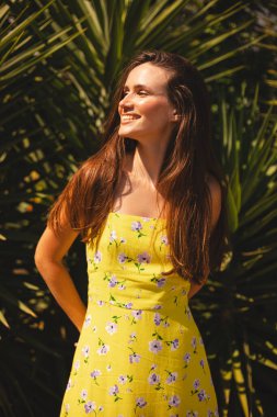 Gülümseyen genç kadın, tropik palmiye ağacının önünde duran tarafa bakıyor. Güneşli bir yaz gününde sarı çiçekli elbise giymek. Neşeli açık hava portresi.