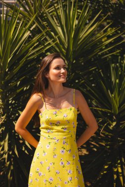 Gülümseyen genç kadın, tropik palmiye ağacının önünde duran tarafa bakıyor. Güneşli bir yaz gününde sarı çiçekli elbise giymek. Neşeli açık hava portresi.