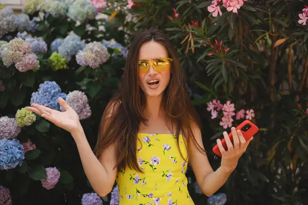 Çiçek fidanlığının yanında cep telefonu kullanan çılgın ve şok edici genç esmer kadın. Mutsuz kadının cep telefonu var. Hayal kırıklığına uğramış üzgün bayan, haberleri dehşete düşürdü. Bu da ne? Olamaz..